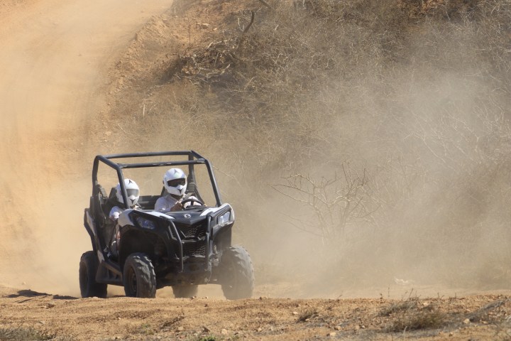 Two people in helmets driving a dune buggy on a dusty off-road trail.