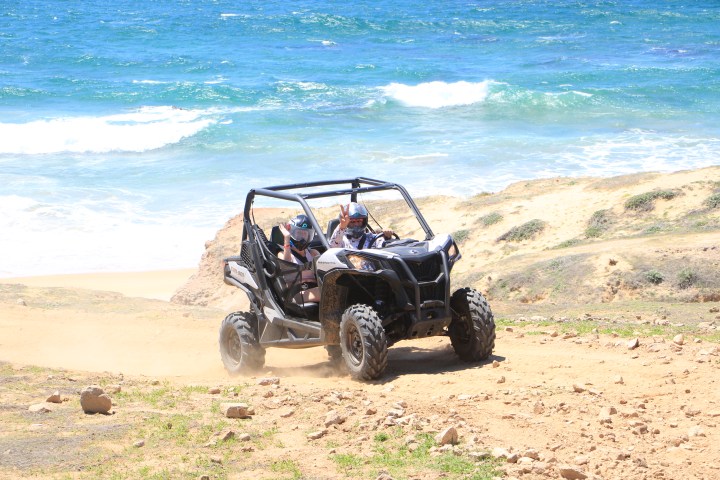 Two people driving a dune buggy on a beach with ocean in background.