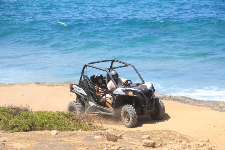 Two people drive a side-by-side ATV on a dirt path near the ocean.