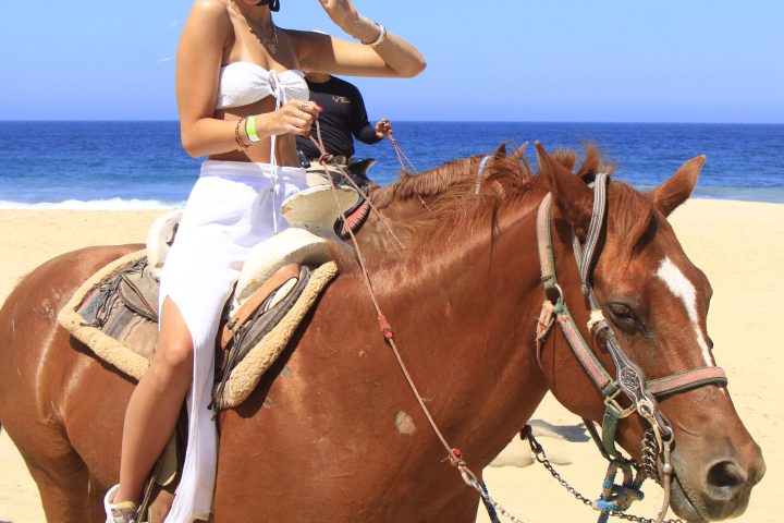 Person in white dress riding a brown horse on a beach, showing a peace sign with the ocean in the background.