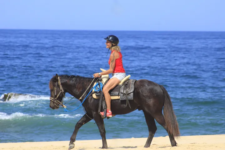 Person in helmet riding a horse on a beach with ocean in the background.