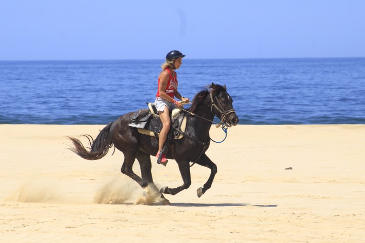 Person in red shirt riding a black horse on a sandy beach near the ocean.
