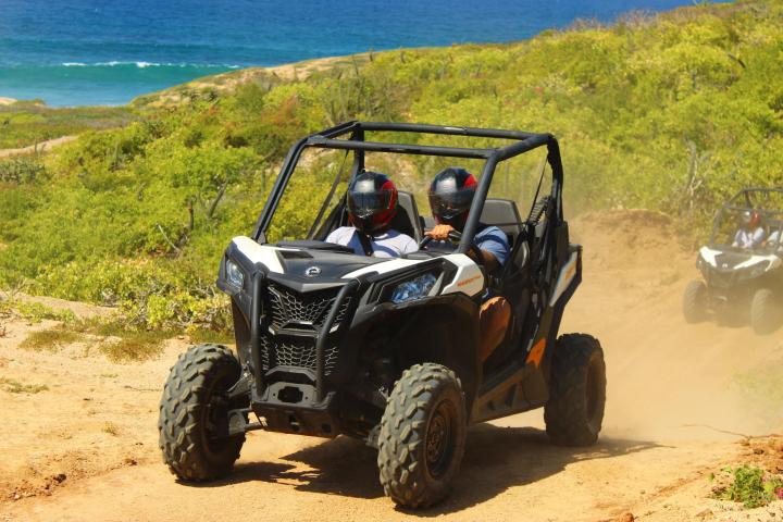 Two people driving an ATV on a dirt path near the ocean, wearing helmets.