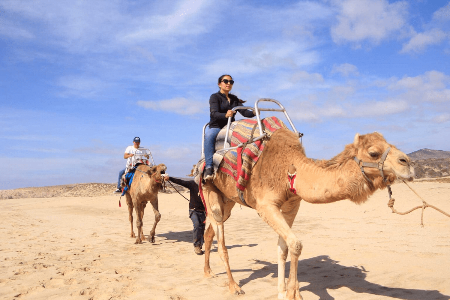 Two people riding camels on a sandy beach under a blue sky.