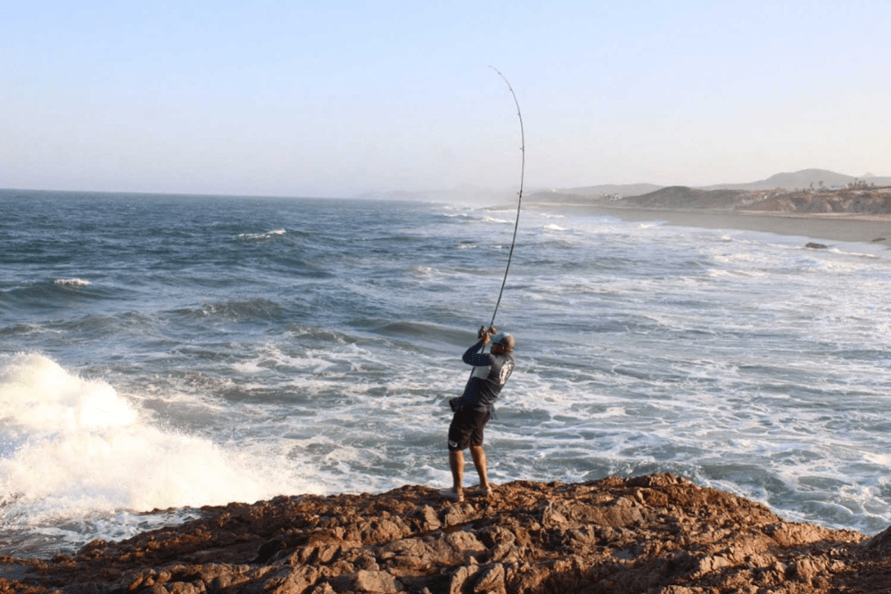 Person fishing from rocky shore with waves in the background.