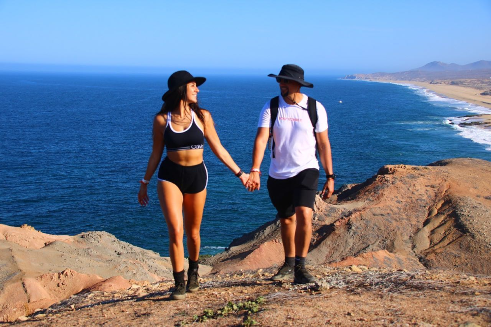 Couple in hats holding hands, walking on cliffside above ocean under clear sky.
