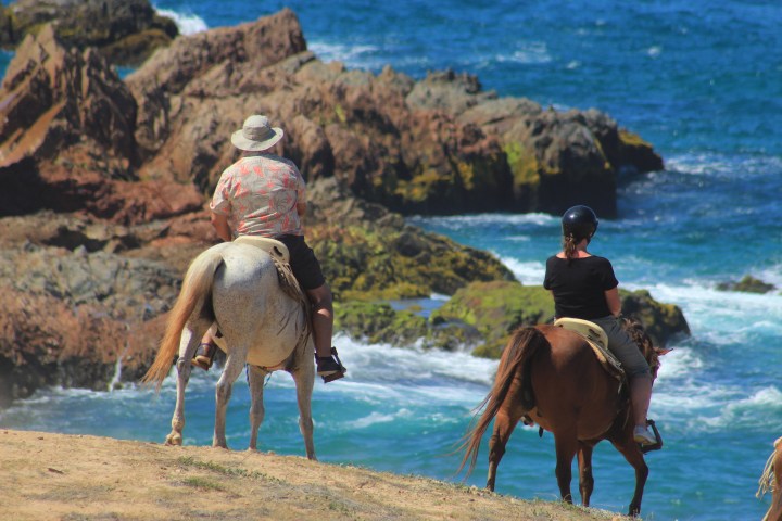 Two people horseback riding near rocky ocean cliffs on a sunny day.