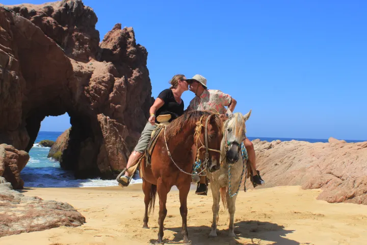 Two people kiss while riding horses on a beach with rock formations and blue sky.