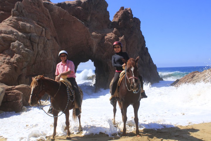 Two people on horseback near rocky arch by ocean waves.